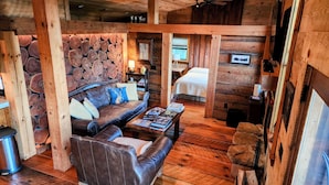 Un acogedor interior de cabaña de madera con un sofá de cuero, una mesa de centro con libros y una cama con edredón blanco.