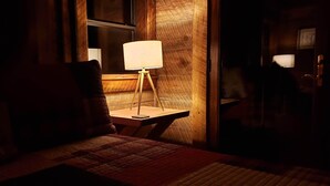 Una habitación tenuemente iluminada, con piso de madera, una pequeña mesa con una lámpara y un sillón con cojín.