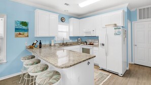 A kitchen with white cabinets, a granite countertop, a refrigerator, and a painting of a beach scene. - ☀️Bikes-Pools-30A-Close to Beach-Chihuahua Flats (Santa Rosa Beach)