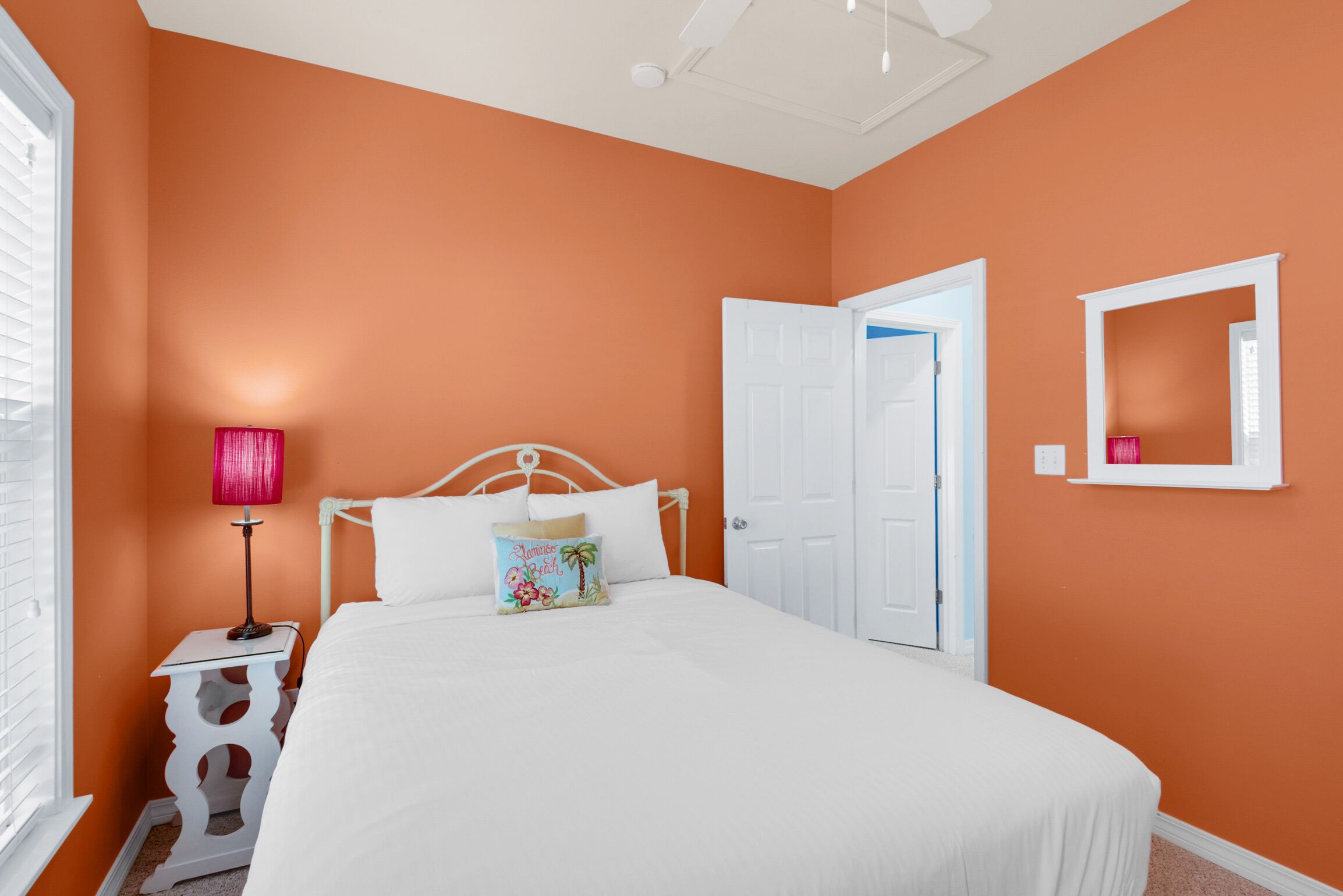 A bedroom with an orange wall, white bed, and a nightstand with a lamp.