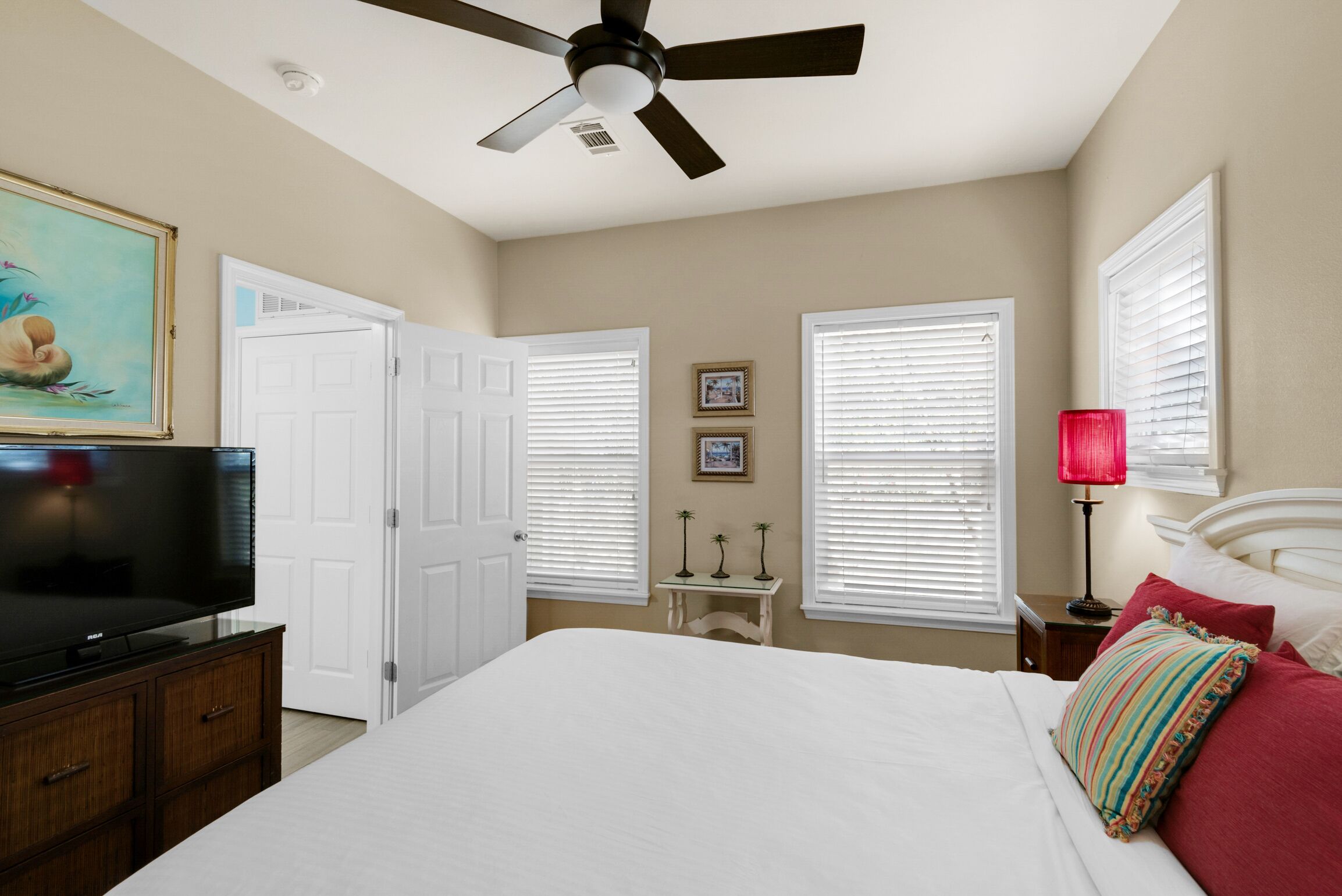 A bedroom with a bed, a ceiling fan, a TV, white walls, and a door.