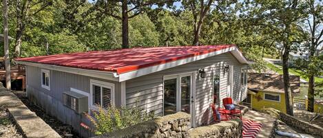 Een klein, gelijkvloers huis met een rood dak, een stenen muur ervoor en een rood-witte strepenmat op de veranda.