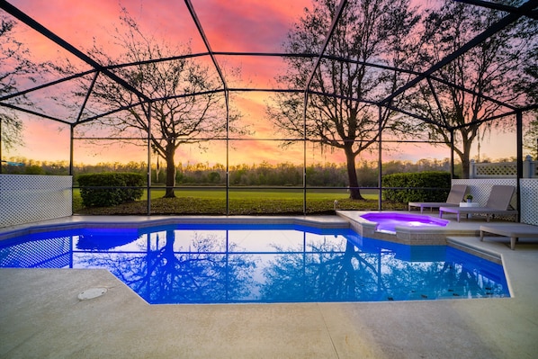 A swimming pool with a glass enclosure, surrounded by lounge chairs and a view of trees and a sunset.