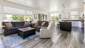 A spacious living room with a dark brown leather sofa, a beige armchair, and a wooden coffee table. A kitchen area with white cabinets and a black island is visible in the background. - Brand new incredible Columbia Falls home - Flathead river walking trail - A/C for hot summer days (Columbia Falls)