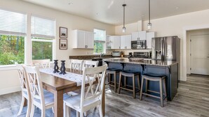A modern kitchen with white cabinets, a stainless steel refrigerator, and a dining area with wooden chairs and a table. - Brand new incredible Columbia Falls home - Flathead river walking trail - A/C for hot summer days (Columbia Falls)