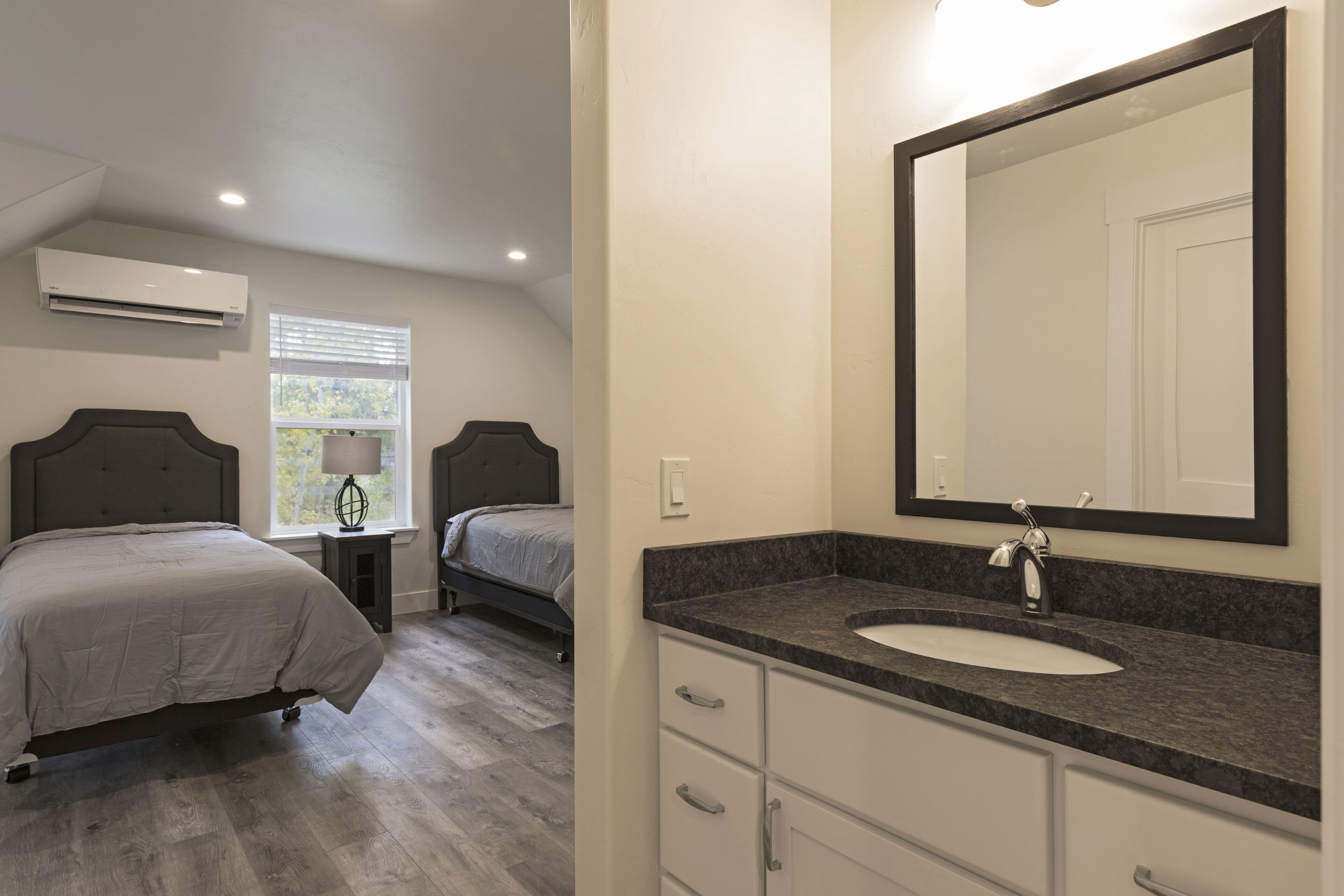 Une chambre avec deux lits, une salle de bain avec un lavabo et un miroir, et un appareil de climatisation.