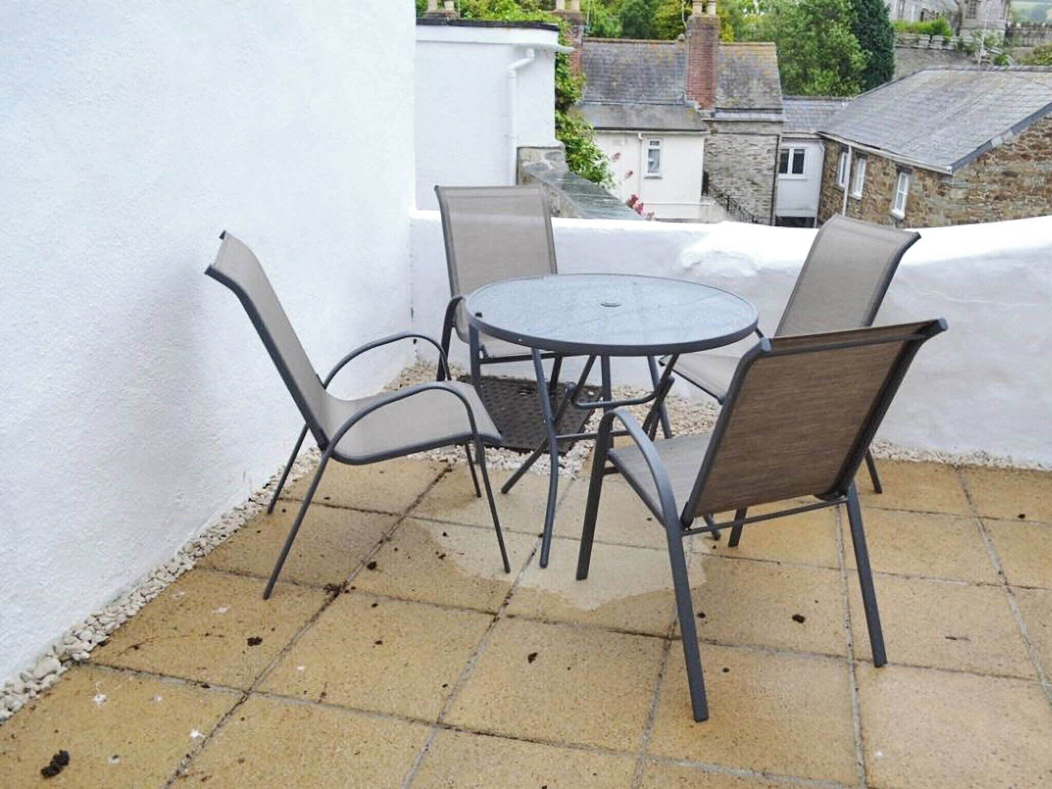 Un pequeño patio al aire libre con una mesa redonda de metal y cuatro sillas, rodeado por un muro blanco y un suelo con baldosas.