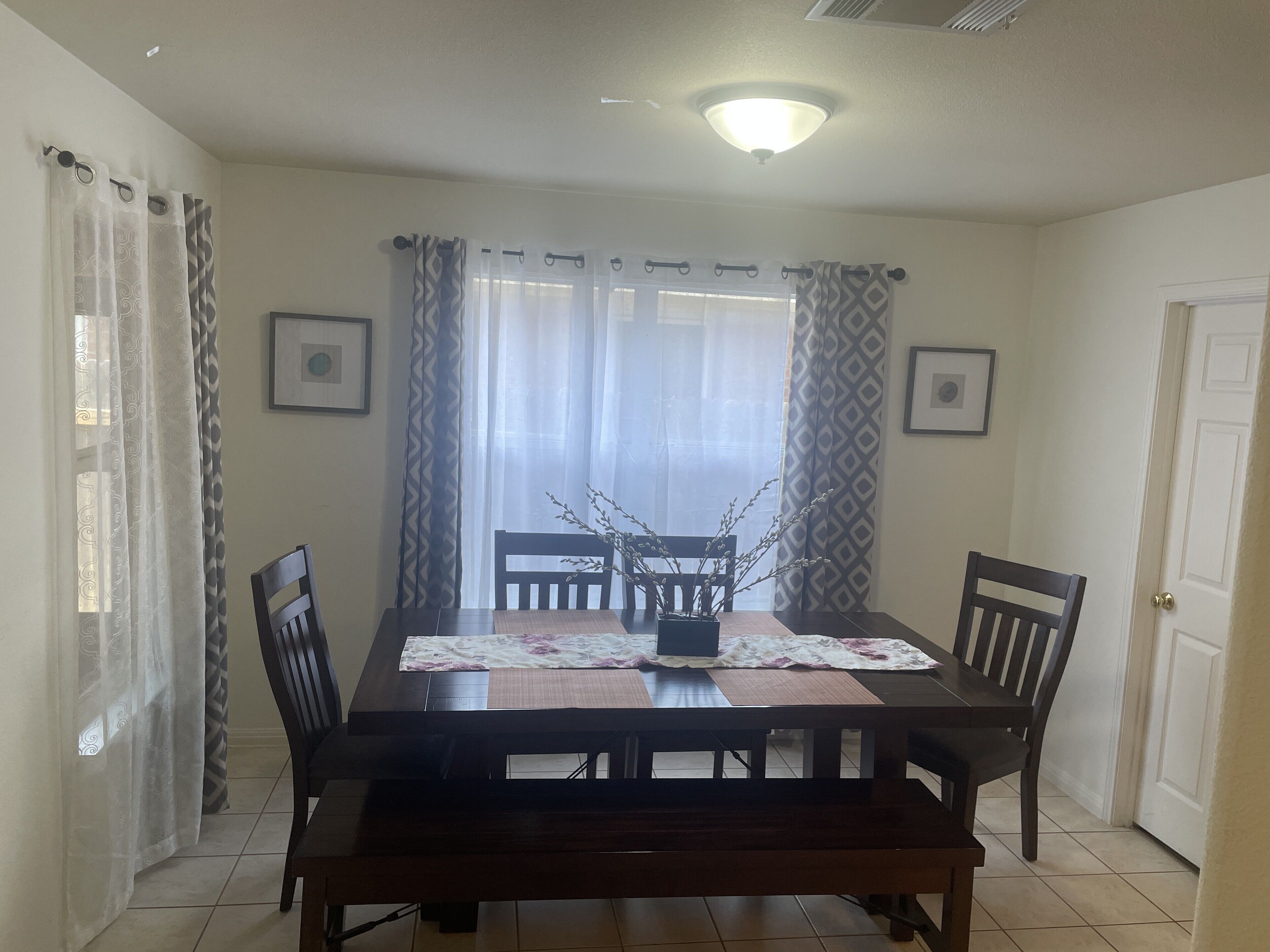 A dining area with a wooden table, chairs, and a bench. There are curtains on the windows, and a ceiling light fixture is visible.