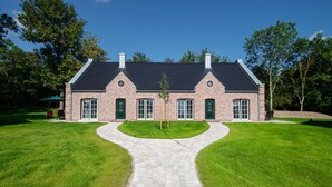 Et murstenshus med en central indkørsel, der fører til hoveddøren, omgivet af velplejede græsplæner.