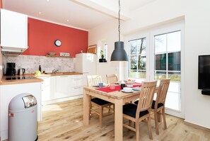 A modern kitchen with white cabinets, a red accent wall, and a dining area with a wooden table and chairs. - Exclusive holiday home with fireplace and sauna, terrace and beach chair, 4 persons (Vitzdorf)
