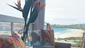 Ein Wohnzimmer mit einem beige-farbenen Sofa, bunten Kissen und einem Glastisch. Ein großes Fenster bietet Blick auf den Strand und den Ozean.