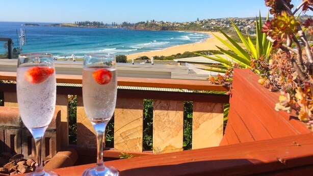 Zwei Gläser mit einem klaren Getränk und Erdbeere als Dekoration auf einem Balkon mit Blick auf einen Strand, Wellen und eine entfernte Stadt.
