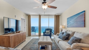 A spacious living room with a flat-screen TV, a wooden entertainment unit, a tufted ottoman, a sofa with plaid cushions, and a painting on the wall. - 🌊Gulf Front Master-2BR Majestic Beach Towers 2-1708🌊 (Panama City Beach)