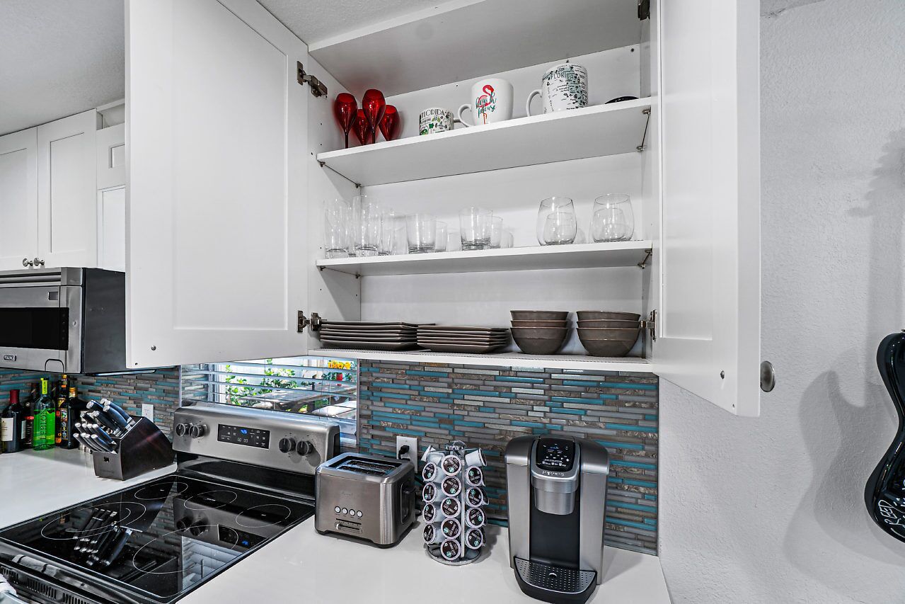 Une cuisine moderne avec des meubles blancs, une cuisinière en acier inoxydable, une machine à café et divers articles en verre disposés sur des étagères.