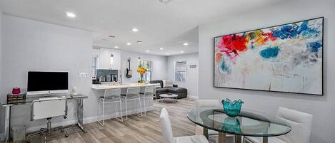 A modern living room with a glass dining table, a sofa, and a kitchen area in the background.