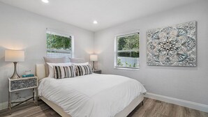 A bedroom with a large bed, two bedside tables, a window with blinds, and a wall-mounted artwork.