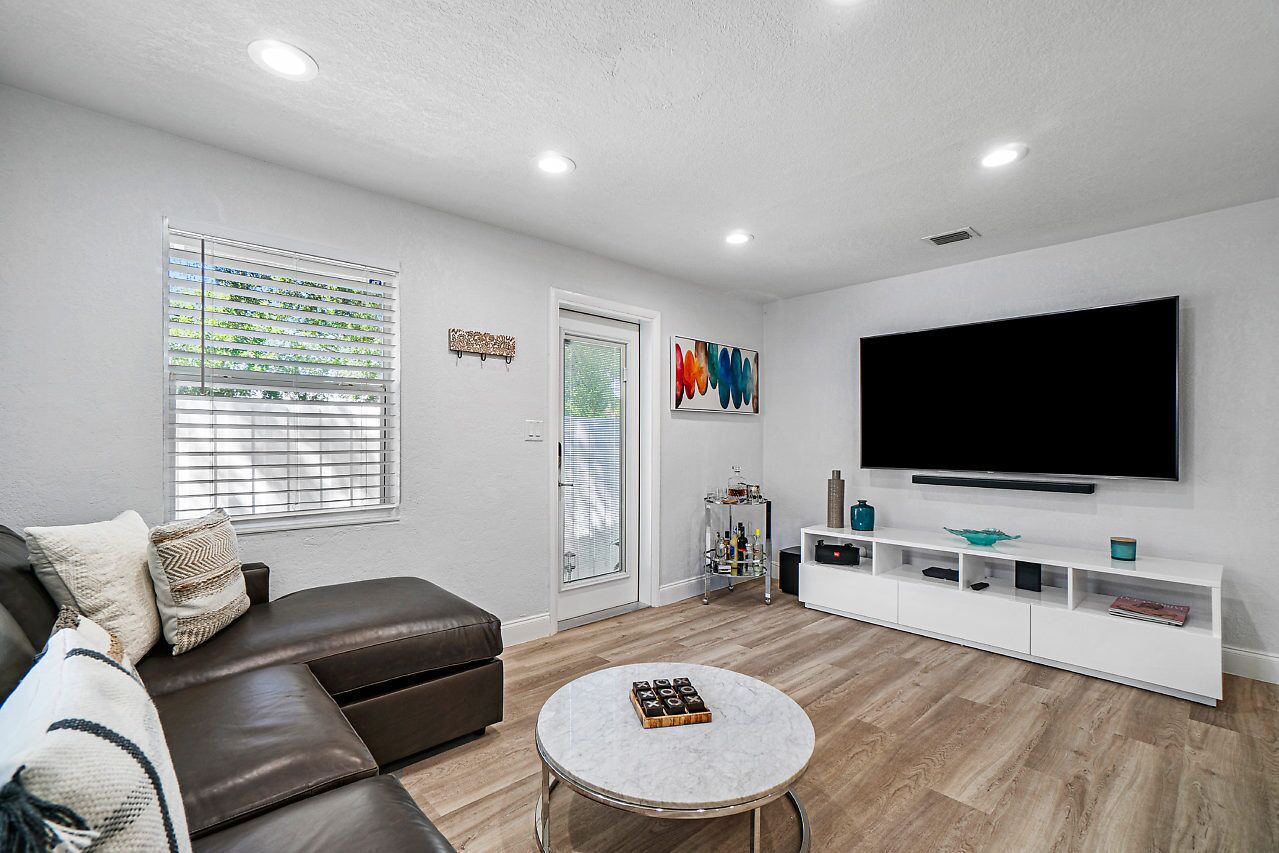 Una sala de estar moderna con un sofá de cuero marrón, una mesa de centro blanca, un televisor de pantalla plana sobre un mueble blanco y una ventana con persianas.