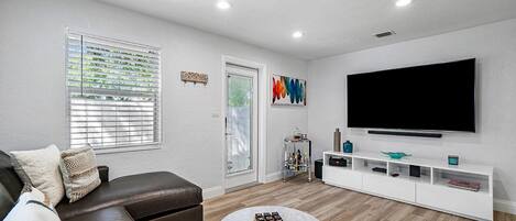 Una sala de estar moderna con un sofá de cuero marrón, una mesa de centro blanca, un televisor de pantalla plana sobre un mueble blanco y una ventana con persianas.