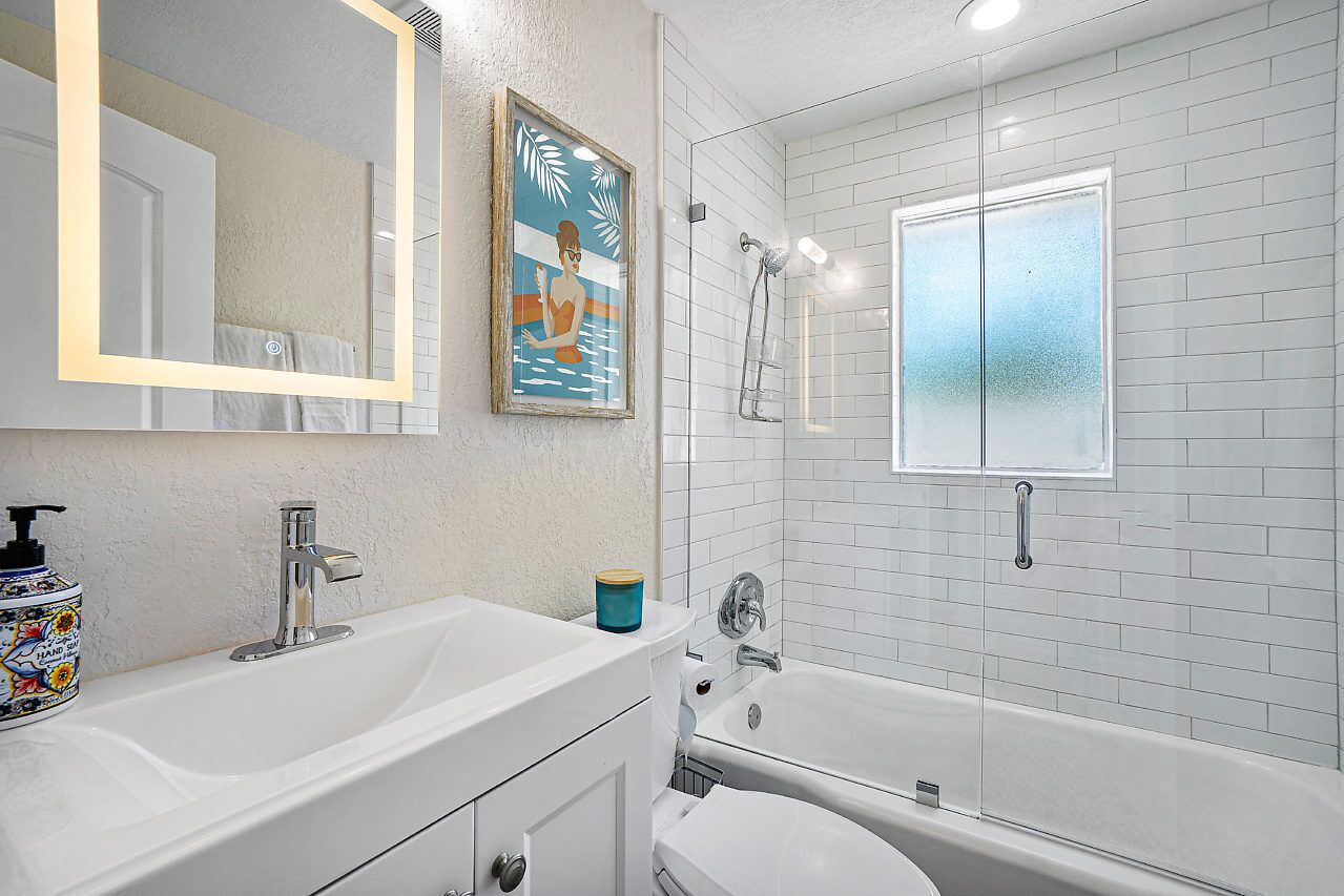 A bathroom with a white sink, a mirror, a framed artwork, a shower, and a window.