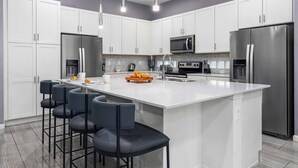 A modern kitchen with white cabinets, a large island with bar stools, stainless steel appliances, and pendant lighting. - Cosmic Home w Space Theme BR, Theater, Arcade, Game Loft, Pool - Mins to Disney (Davenport)
