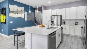 A modern kitchen with white cabinets, a stainless steel refrigerator, and a central island with a sink. - Cosmic Home w Space Theme BR, Theater, Arcade, Game Loft, Pool - Mins to Disney (Davenport)