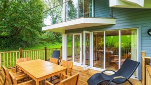 Een houten terras met een tafel en stoelen, een glazen deur die leidt naar een buitenruimte, en een blauw huis op de achtergrond.