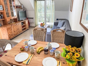 A dining area with a wooden table set for two, surrounded by wicker chairs. A television is mounted on the wall, and a sofa is visible in the background.