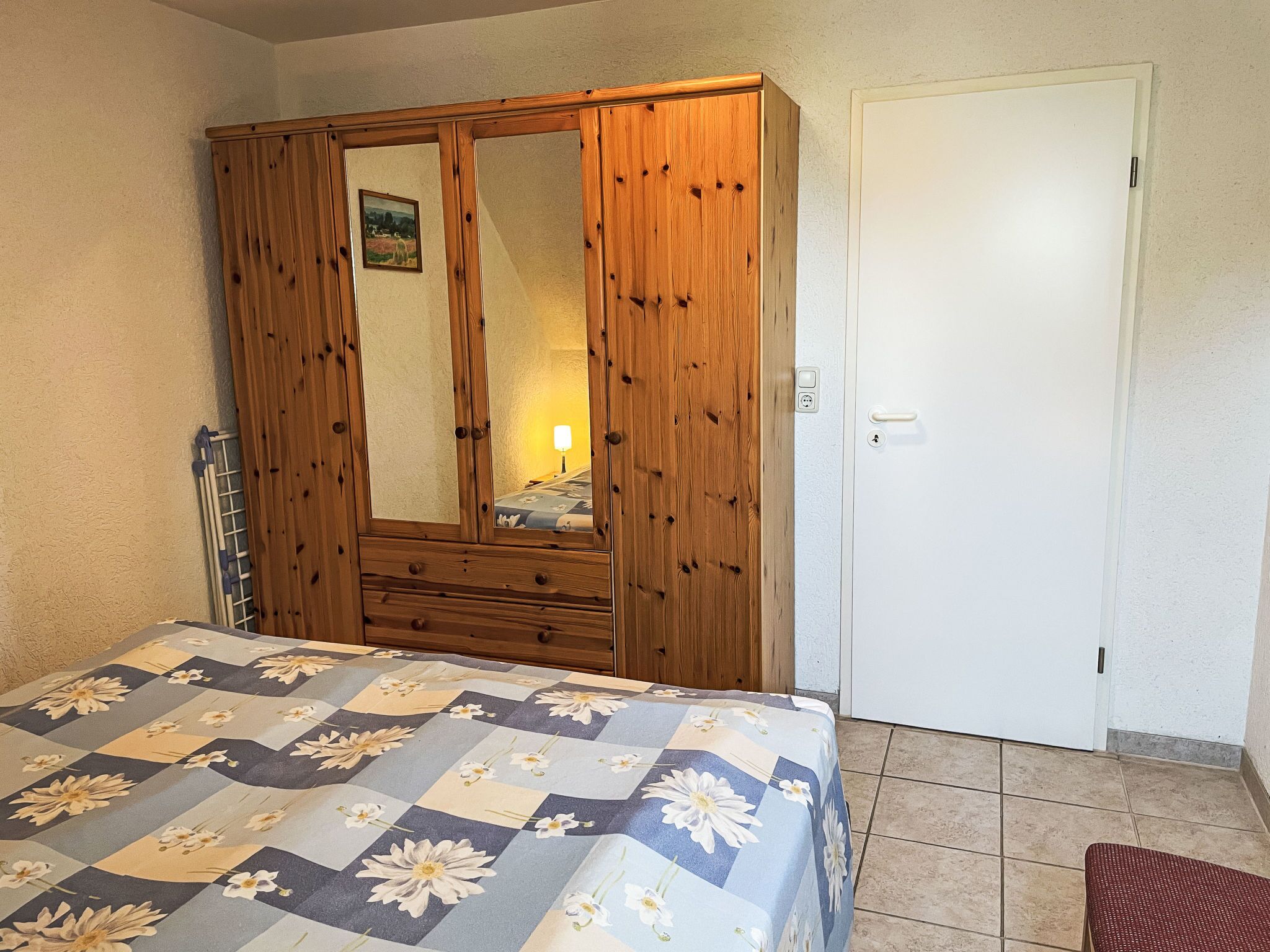 A bedroom with a wooden wardrobe, a bed with a floral bedspread, and a white door.