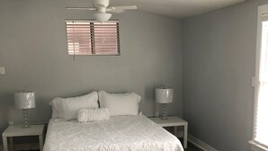 Un dormitorio con cama, dos mesitas de noche, ventilador de techo, ventana con persianas y puerta.