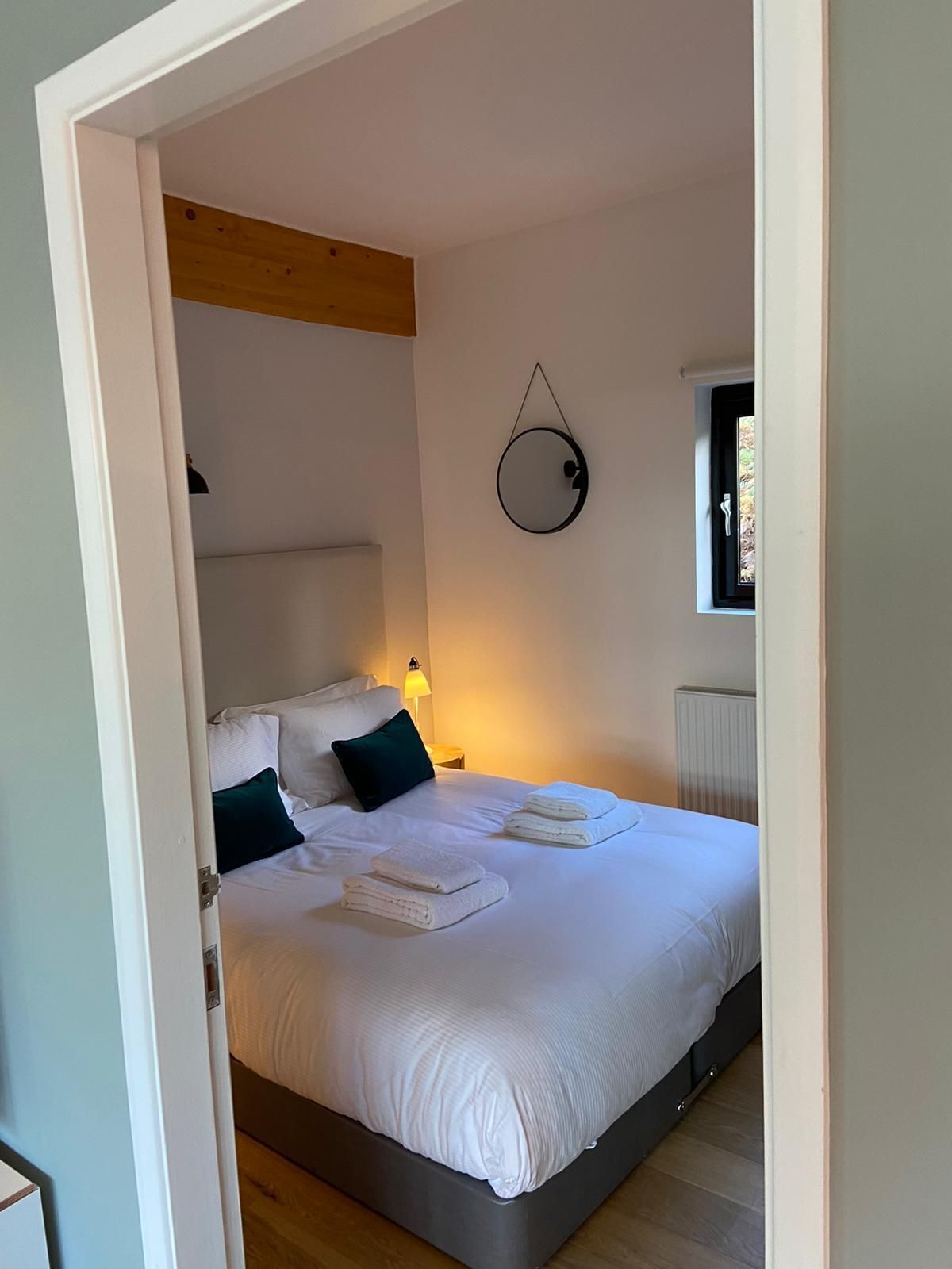 A bedroom with a bed, pillows, a bedside lamp, a window, and a wall-mounted circular mirror.