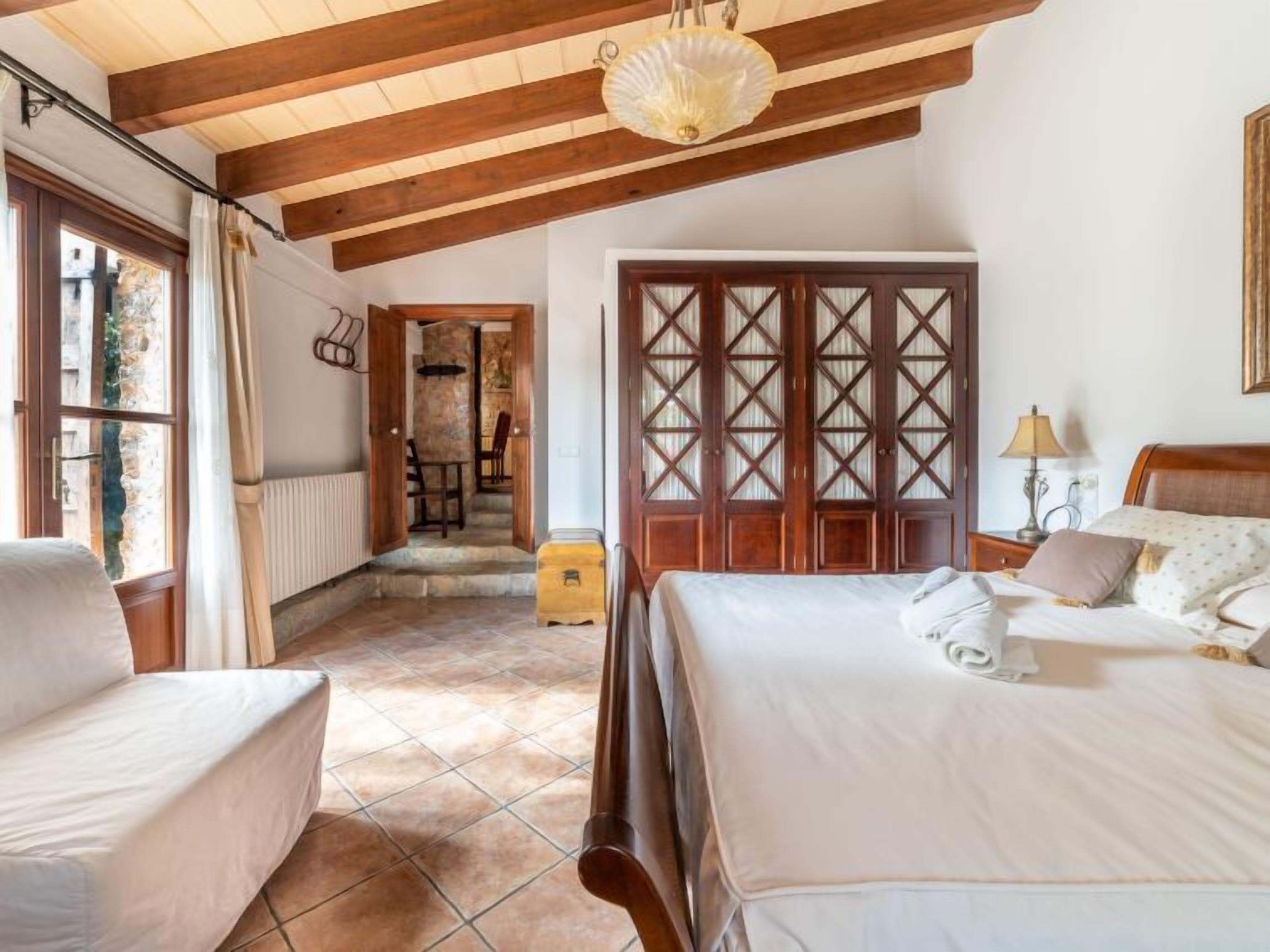A bedroom with two beds, wooden beams, a stone wall, and a view of the outside.