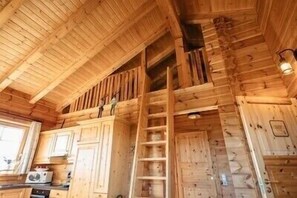 L’intérieur d’une cabane en bois comprend un espace de couchage en mezzanine, une cuisine avec un poêle et une échelle menant à la mezzanine.