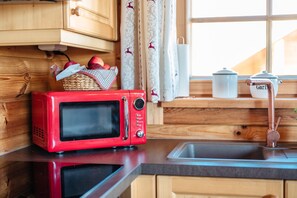 Une cuisine avec des meubles de rangement en bois, un micro-ondes rouge, un évier et une fenêtre avec des rideaux.