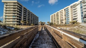 Ein hölzerner Boardwalk führt zu zwei Hochhäusern mit zahlreichen Balkonen, umgeben von Palmen und sandigem Gelände.