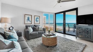 Ein modernes Wohnzimmer mit einem grauen Sofa, einem runden gewebten Couchtisch und einem Deckenventilator. An den Wänden hängen gerahmte Pflanzenprints, eine Lampe und ein Blick auf das Meer durch eine Schiebetür.