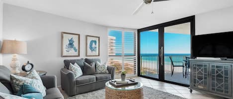 A modern living room with a grey sofa, a round woven coffee table, and a ceiling fan. There are framed botanical prints on the wall, a lamp, and a view of the ocean through a sliding glass door.