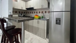 A kitchen with light wood cabinets, a stainless steel refrigerator, a granite countertop, and a dining area with two chairs.