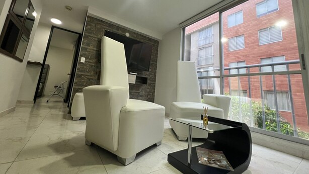 A modern living room with a sofa, armchair, and coffee table, a flat-screen TV mounted on a stone wall, and a large window with a view of a brick building.