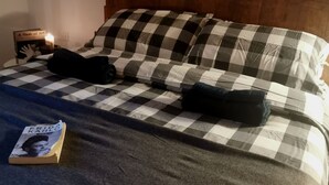 Uma cama com colcha xadrez, duas almofadas, um livro e uma mesa de cabeceira com uma vela.