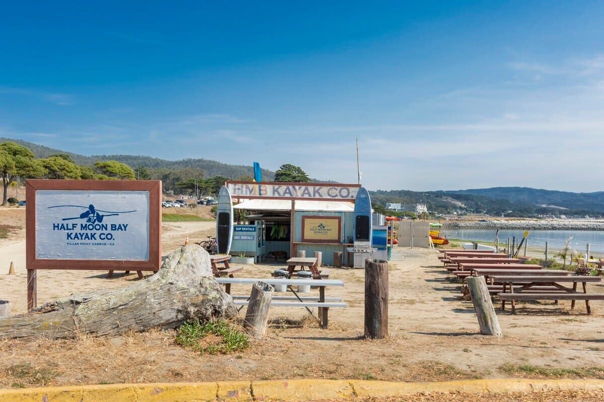 Un point de location de kayaks situé au bord de la plage, avec une enseigne indiquant « Half Moon Bay Kayak Co. », des tables de pique-nique et une vue sur l’océan.
