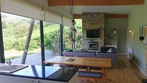 A modern kitchen with a large window, a dining area with a table and chairs, a stone fireplace, and a TV mounted on the wall.