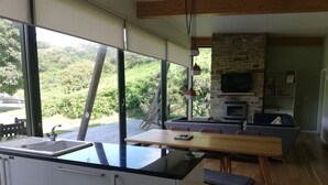 A modern kitchen with a large island, dining area with a wooden table and chairs, a stone fireplace, and a view of greenery through large windows.