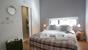 A modern bedroom with a plaid bedspread, a green ottoman, a bedside table with a lamp, and a view through a window.