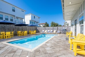 Zwembad met betegelde vloer, omringd door gele stoelen en parasols, met een houten omheining en gebouwen op de achtergrond.