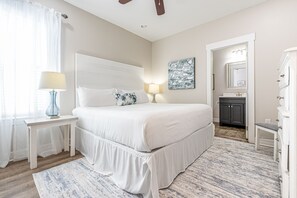 Een slaapkamer met een groot bed, nachtlampjes, een nachtkastje, een raam met dunne gordijnen, een schilderij aan de muur en een badkamer zichtbaar door een geopende deur.