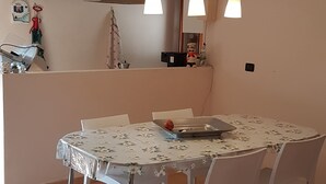 Ein Essbereich mit einem runden Tisch, der mit einer blumigen Tischdecke bedeckt ist. Weiße Stühle stehen darum. Über dem Tisch befinden sich hängende Pendelleuchten.