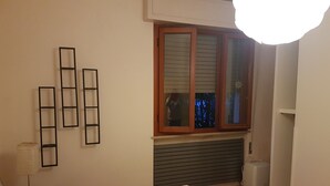 Ein Schlafzimmer mit Bett, einem Fenster mit Jalousien, einer Heizung, einem kleinen Tisch mit Lampe und einem an der Wand befestigten Regal.