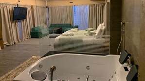 Habitación de hotel moderna con una bañera grande, un sofá verde, un televisor de pantalla plana y un amplio ventanal con cortinas.