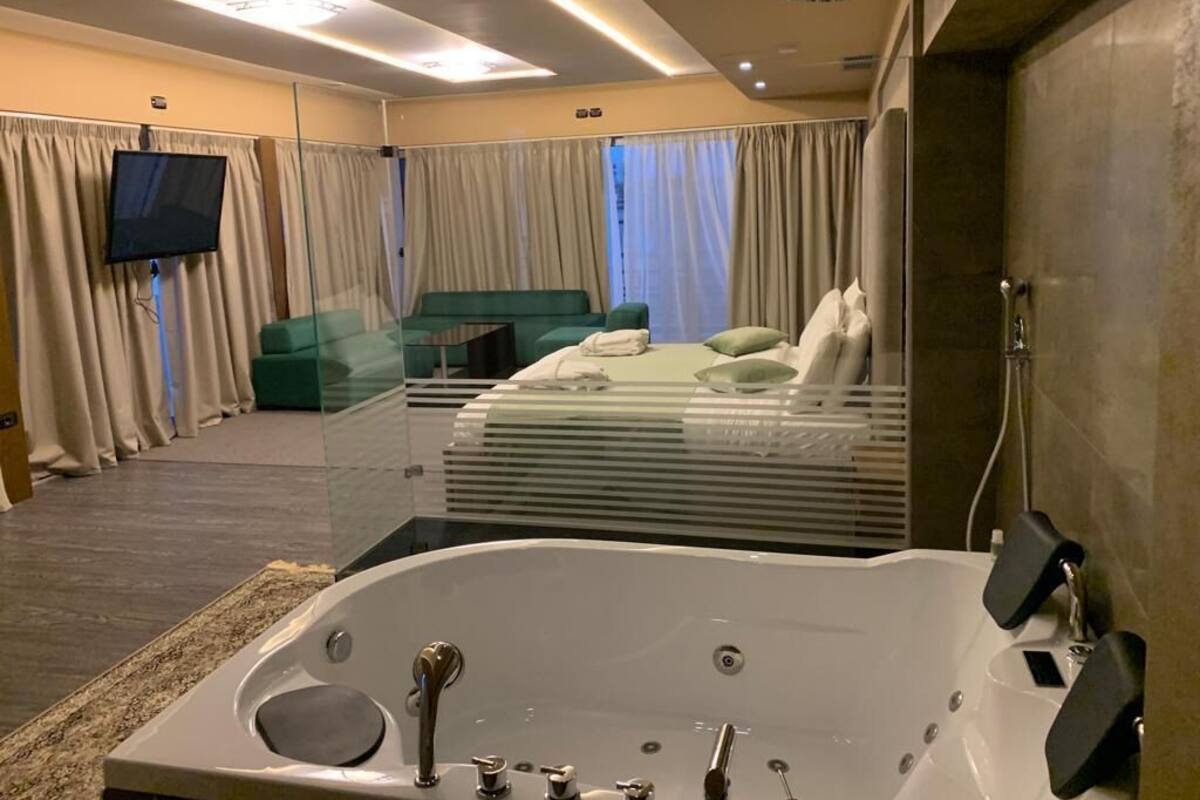 A modern hotel room with a large bathtub, a green sofa, a flat-screen TV, and a large window with curtains.