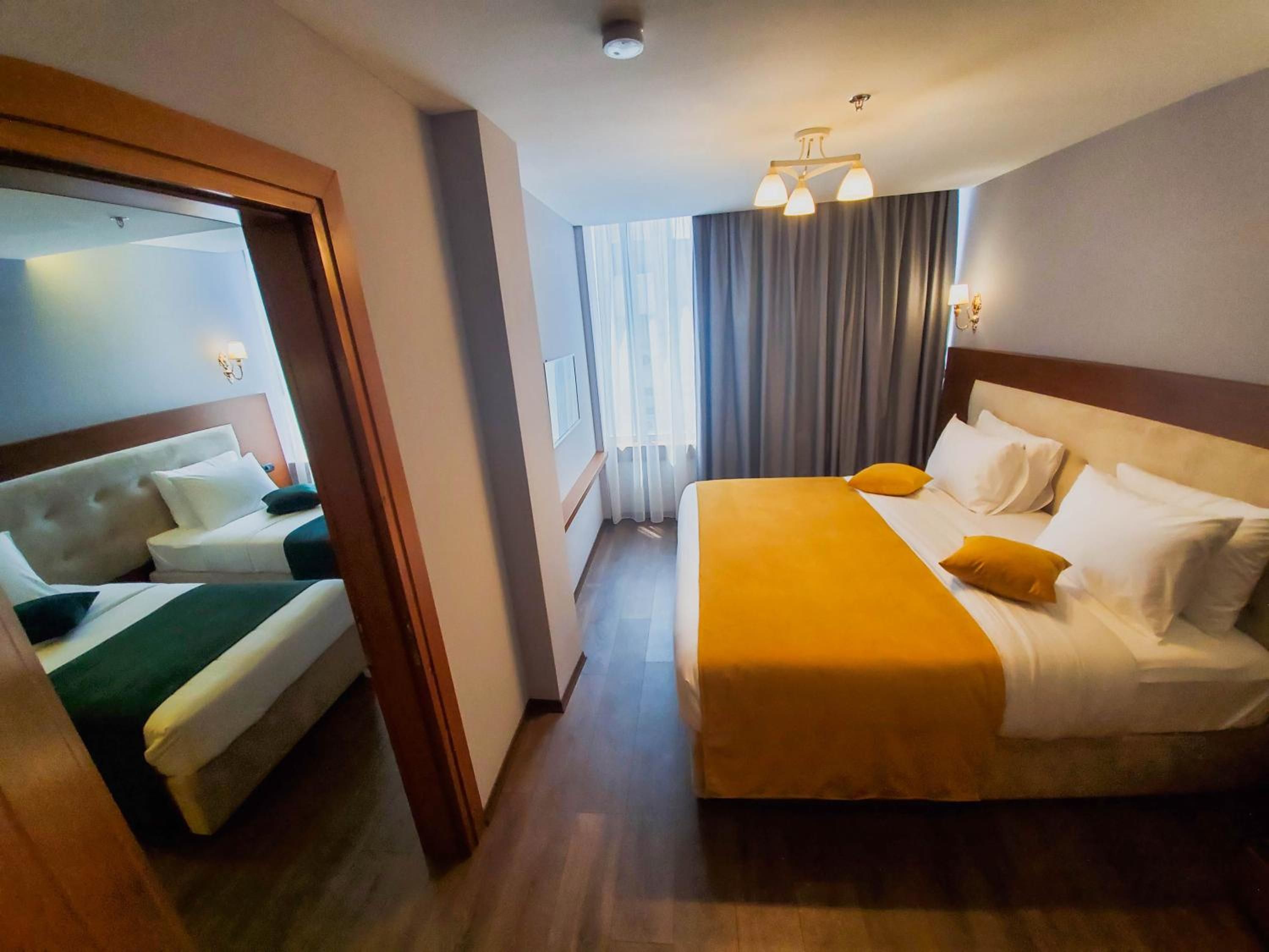 A hotel room with a large bed, a smaller bed, a wooden floor, a window with curtains, and a ceiling light fixture.